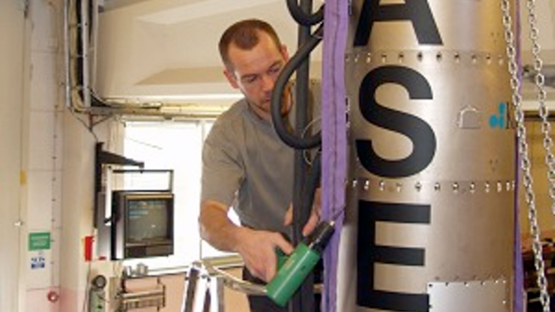 a man in a gray shirt holds a green and black tool as he works on a metal rocket with larger black lettering