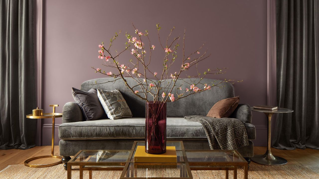 A purple painted living room with a gray couch and gray curtains and two mismatched gold side tables and coffee table. A large pink floral plant is in a vase on the coffee table