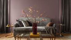 A purple painted living room with a gray couch and gray curtains and two mismatched gold side tables and coffee table. A large pink floral plant is in a vase on the coffee table