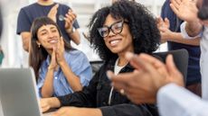 An office worker smiles as her coworkers gather around and applaud.