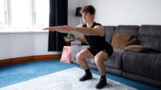 man performing a squat with his hips lowered close to a sofa and arms outstretched in front, in a living room setting