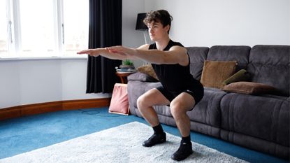 man performing a squat with his hips lowered close to a sofa and arms outstretched in front, in a living room setting 