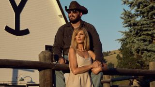 Cole Hauser as Rip Wheeler sitting on a fence and wearing sunglasses and a cowboy hat with Kelly Reilly as Beth Dutton standing below him holding onto his right leg.