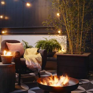 Seating area in garden with a fire pit on tiled floor, a bamboo tree in a pot, and festoon lights hanging from the fence