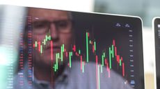 A man's face is reflected in a computer screen showing volatile stock movement.