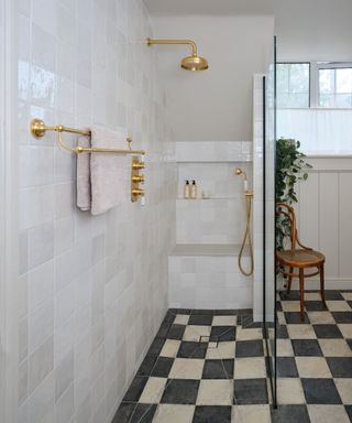 A wet room with glossy white square tiles on the walls and black and white checkerboard tiles on the floor. The shower area features brass fixtures, including a rainfall showerhead and a handheld shower, along with a wall-mounted towel rail. A built-in bench is set into the shower space.