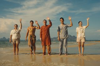 Five members of a hotel staff dressed in different uniforms wave smilingly at a boat approaching out of camera while standing on a tropical beach.