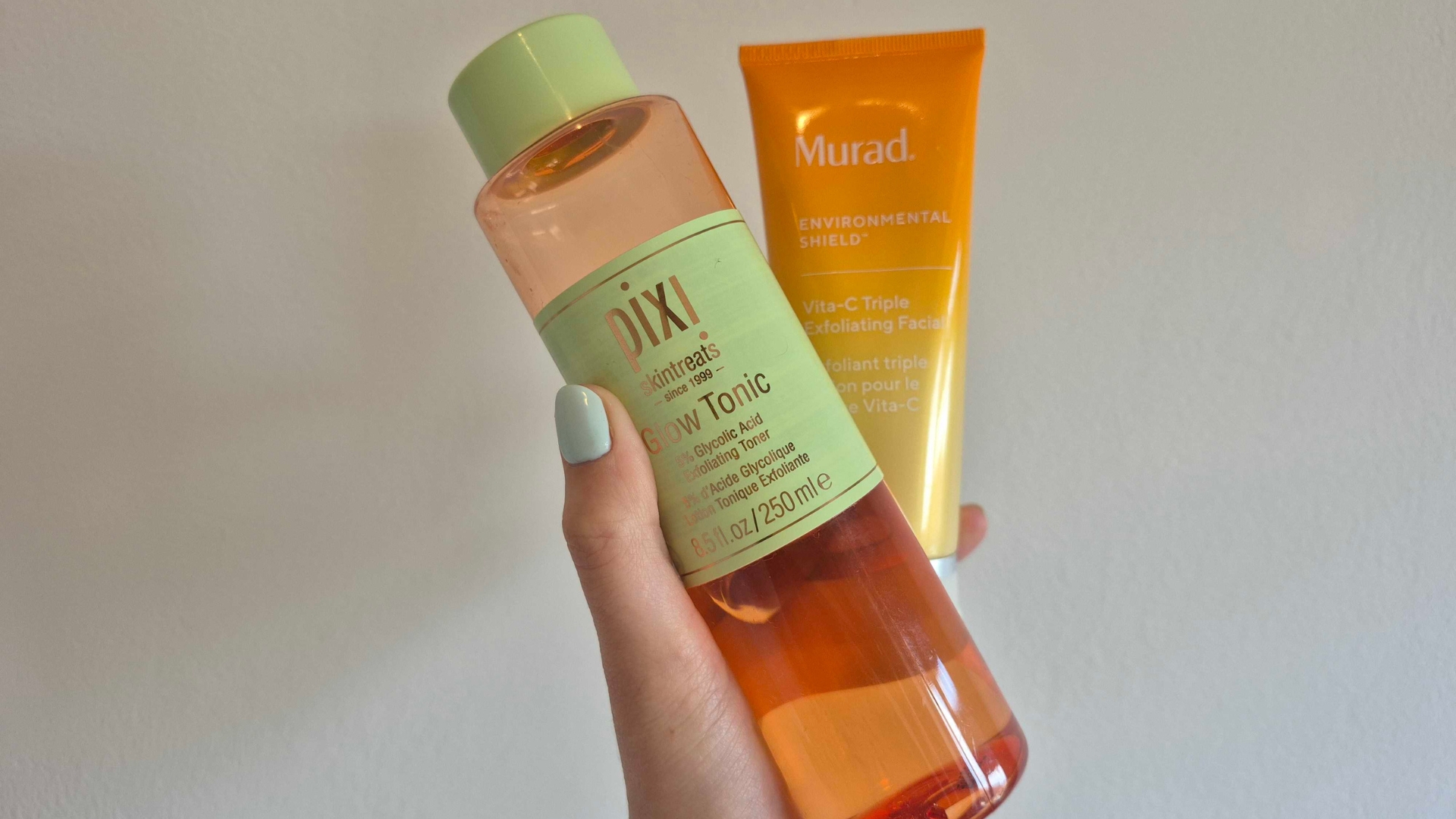 Digital Beauty Writer, Naomi Jamieson's hand is pictured holding two of the Best facial exfoliators featured in this guide: the Pixi Glow Tonic and Murad Vita-C Triple Exfoliating Facial, in front of a white wall