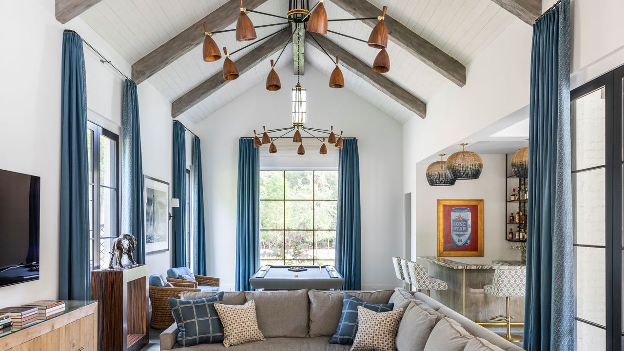 Example of wood ceiling ideas. Large living room with vaulted ceiling, dark wood beams and white wooden paneled ceiling, blue curtains, gray corner sofa, pool table, wooden unit with tv mounted above, two copper chandeliers