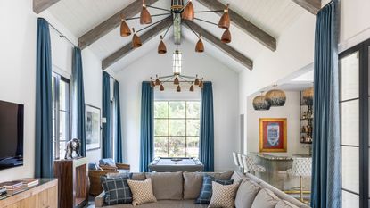 Example of wood ceiling ideas. Large living room with vaulted ceiling, dark wood beams and white wooden paneled ceiling, blue curtains, gray coer sofa, pool table, wooden unit with tv mounted above, two copper chandeliers