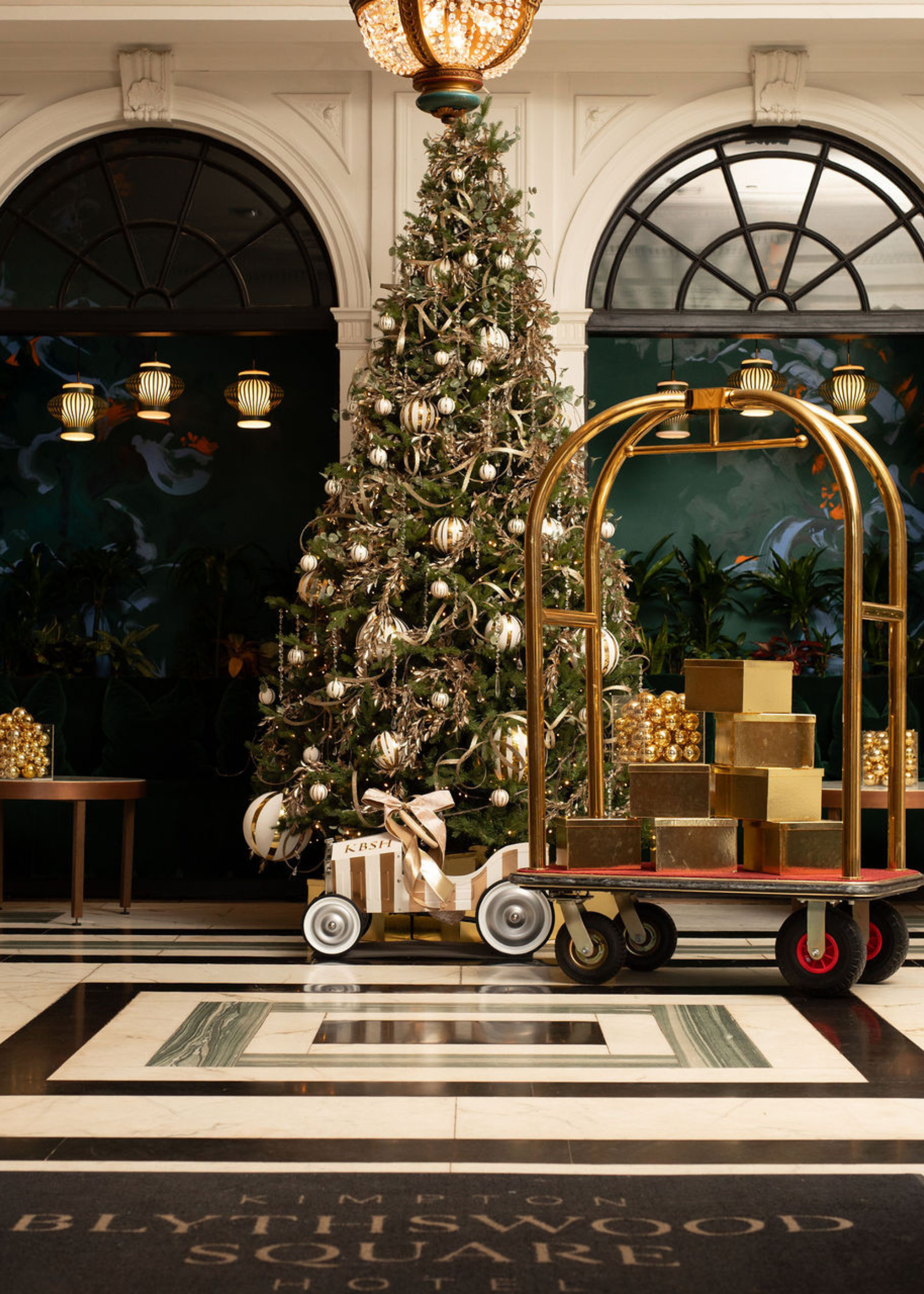 A large Christmas tree in a hotel lobby beside a golden lobby trolley with gifts on top. Behind the tree are archways leading to different parts of the hotel.