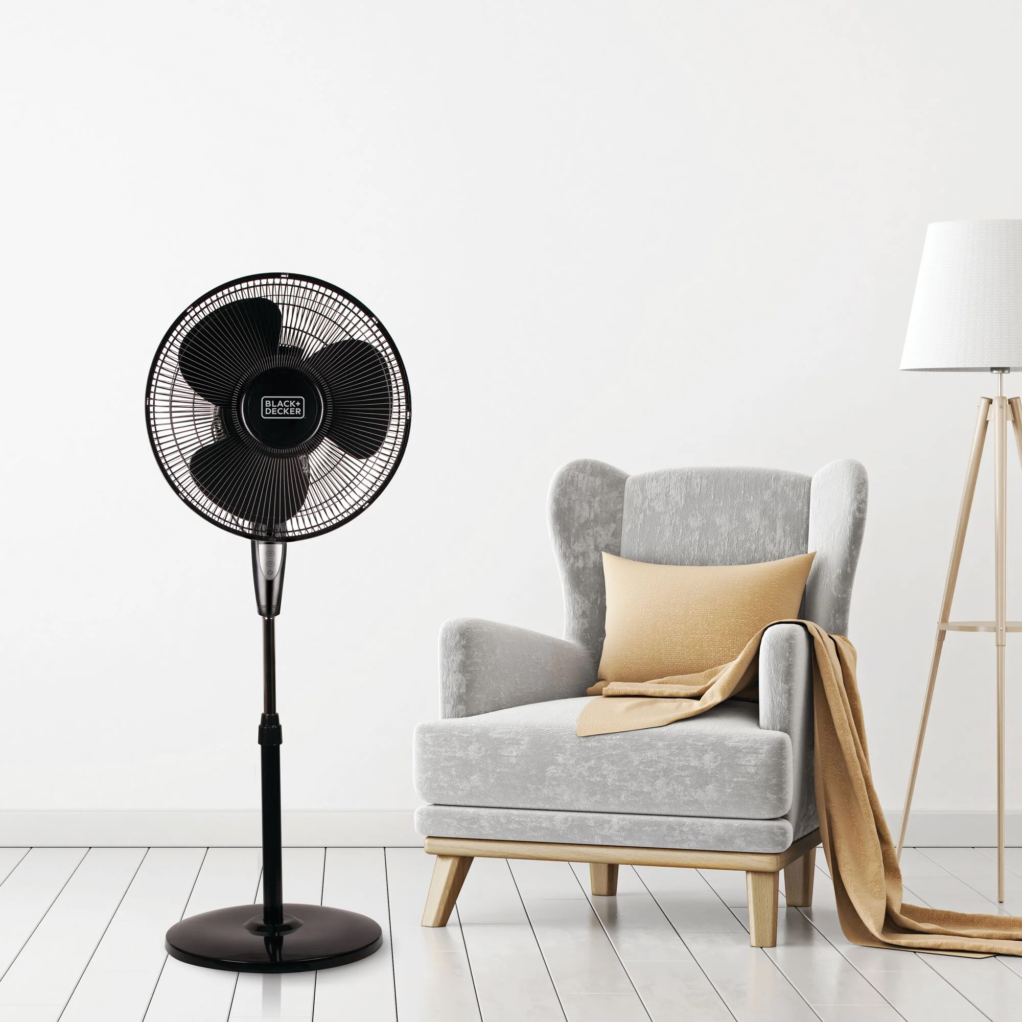 A black BLACK+DECKER pedestal fan in a white room with a grey armchair and floor lamp