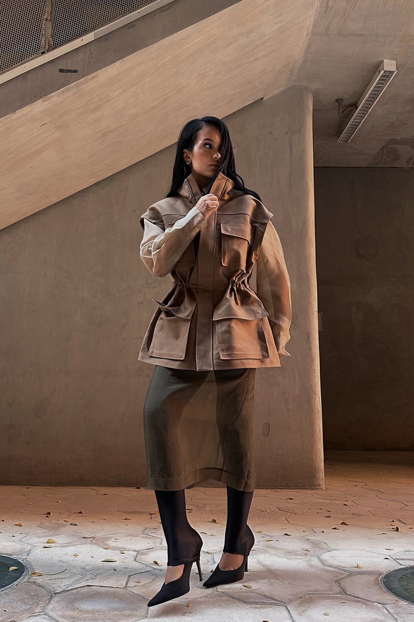 Layering staples for spring 2026 is shown in a street style photo with a woman standing outside while wearing a tan cargo jacket layered on top of an olive green sheer skirt with black leggings styled underneath and worn with black pointed pumps