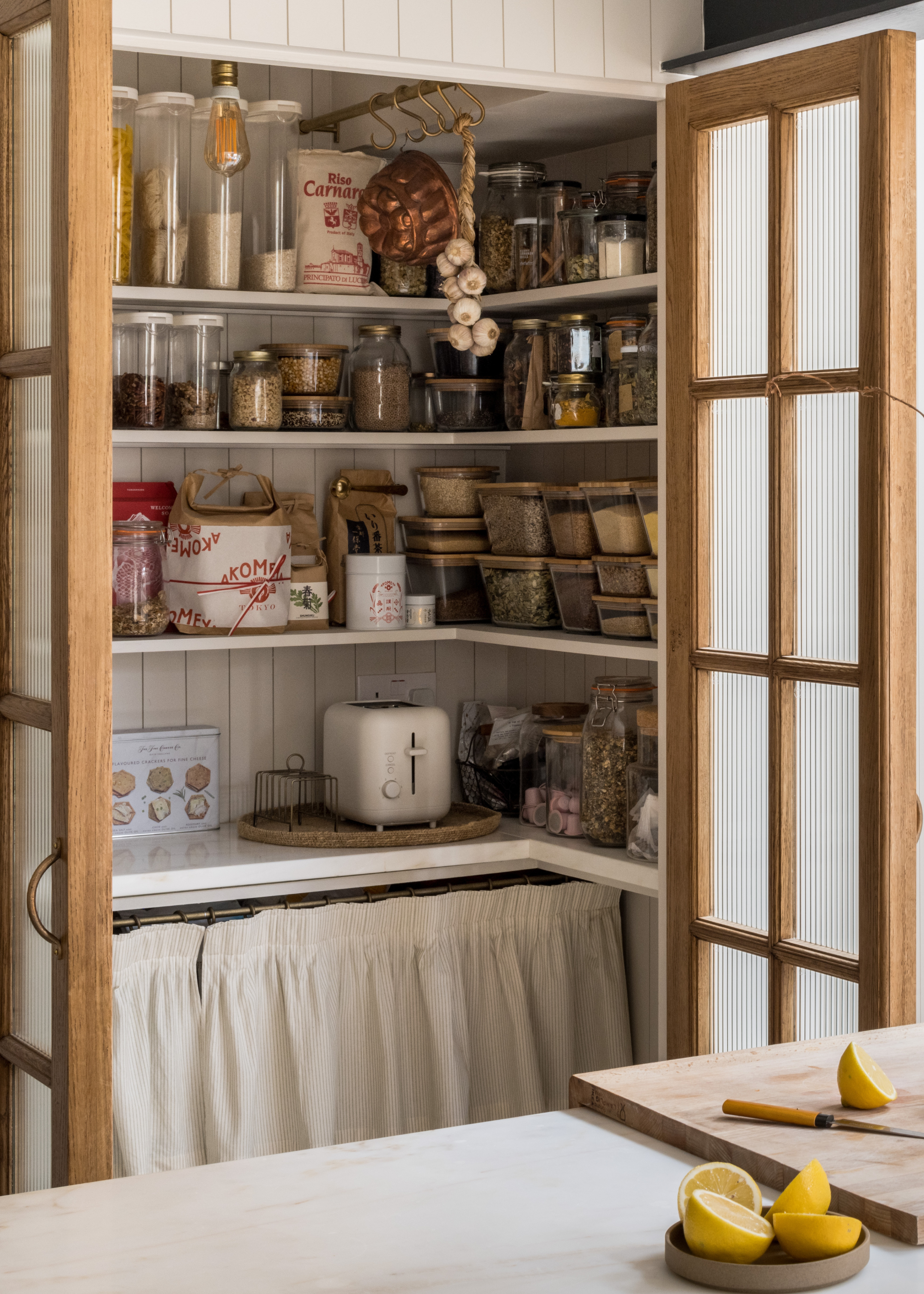 A pantry with glass bottles and containers, hanging garlic, condiments, a bowl of sliced lemons, a chopping board, an oven, and ingredients