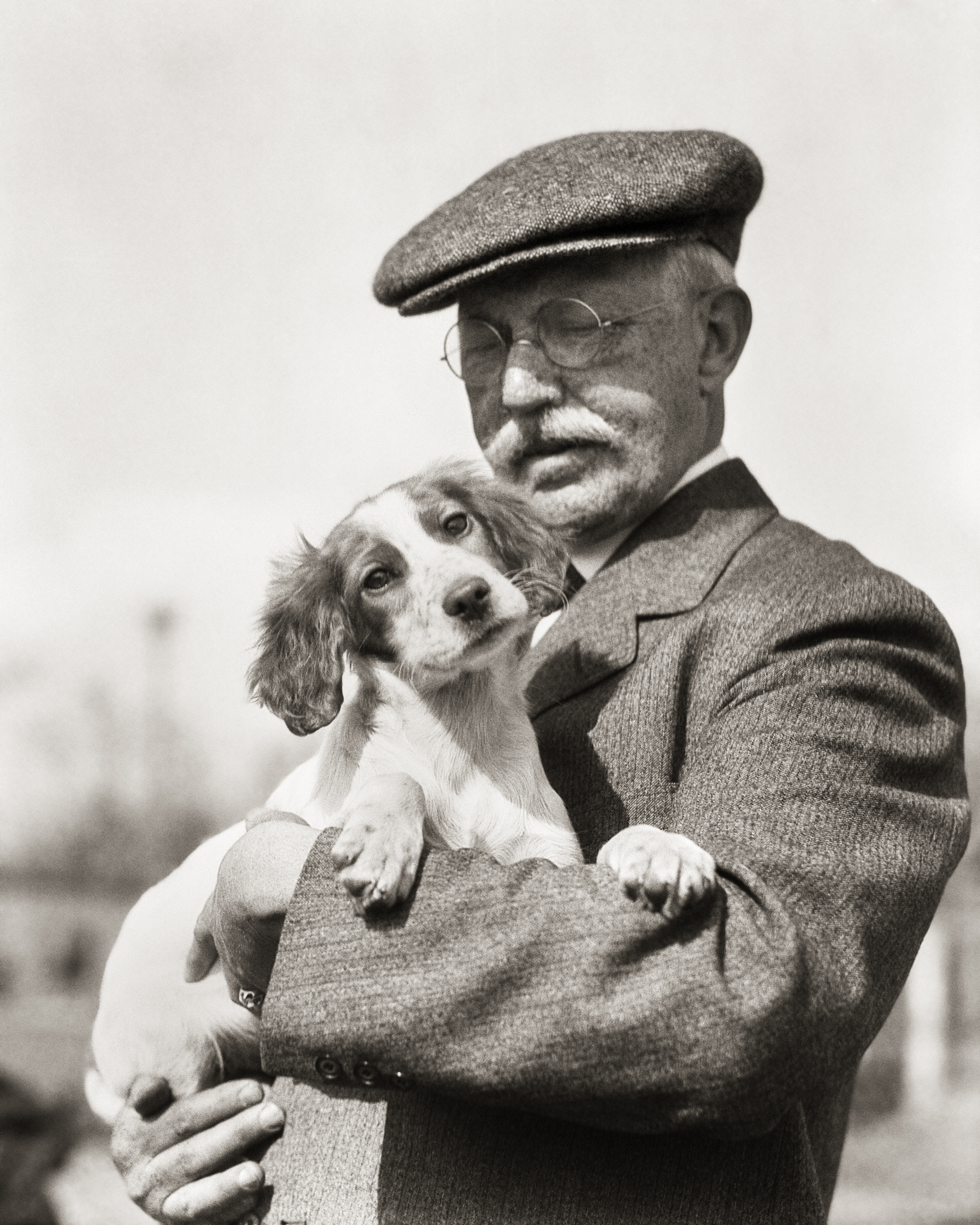 Vintage black-and-white photograph of a man in a flat cap and glasses holding an Irish Red and White Setter puppy in his arms.
