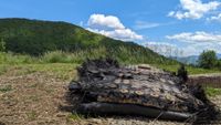 a large black piece of fiberglass covered in metal bolts and plates lies on the ground beside a trail leading into a forest. mountains can be seen rolling in the distance