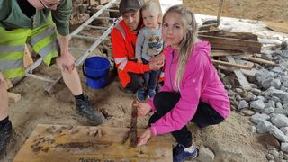 A woman in a pink coat holds a rusty sword next to her toddler son and husband, who is wearing a red jacket. They are in a construction area next to an archaeologist in a green shirt.