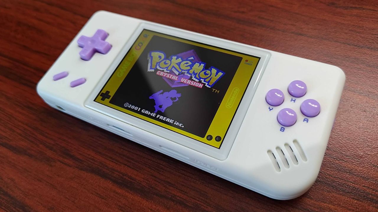 Anbernic RG28XX handheld with Pokemon Crystal on screen sitting on woodgrain desk.