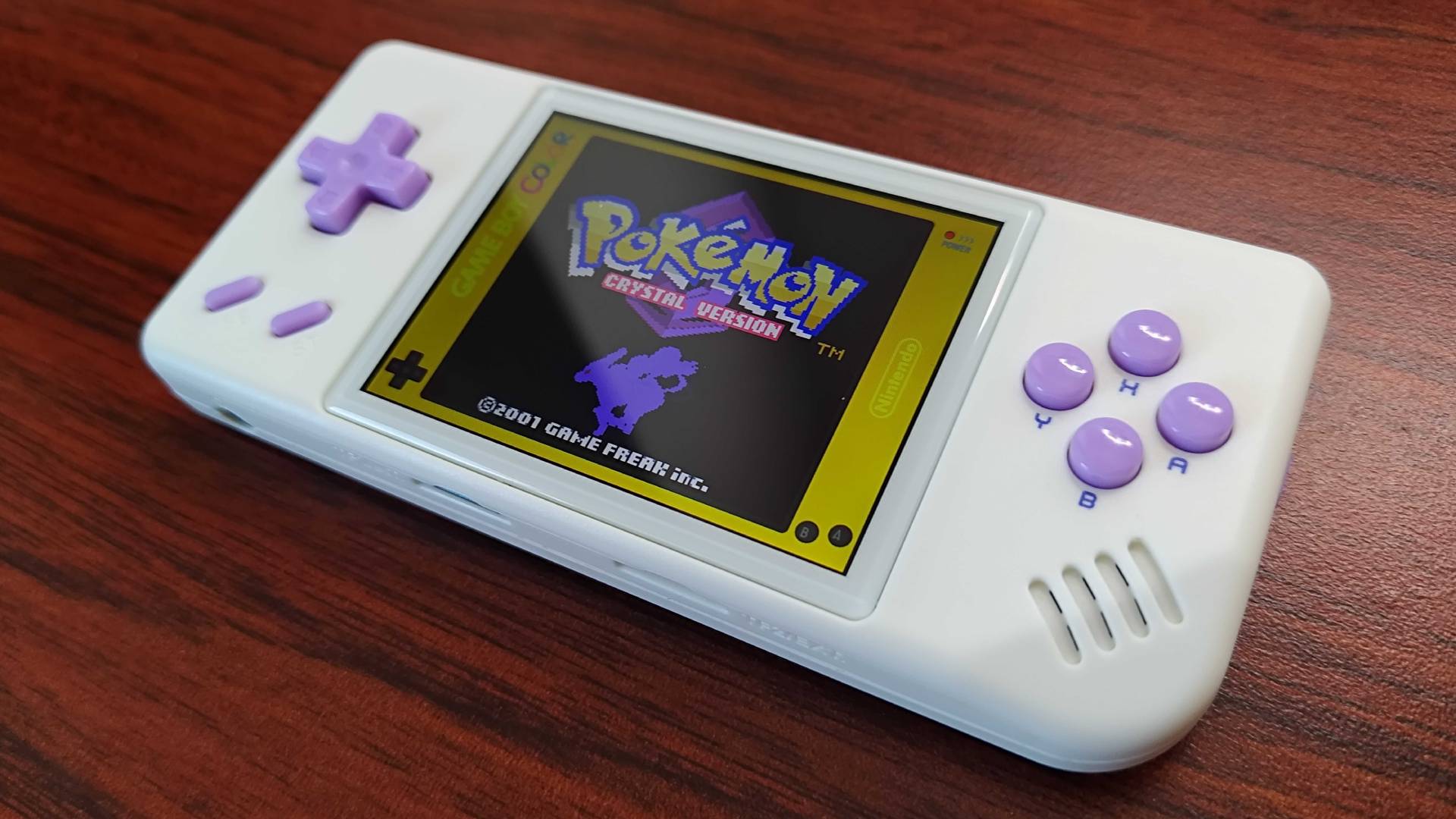 Anbernic RG28XX handheld with Pokemon Crystal on screen sitting on woodgrain desk.