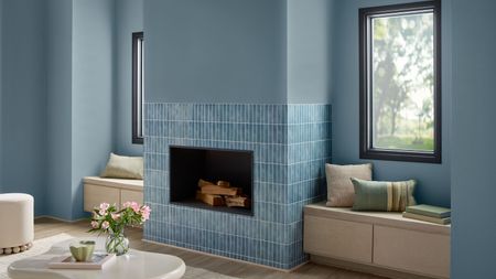 A living room painted powder blue, with a blue-tiled fireplace, two wooden window seats, and a curved white coffee table with a vase of flowers on top.