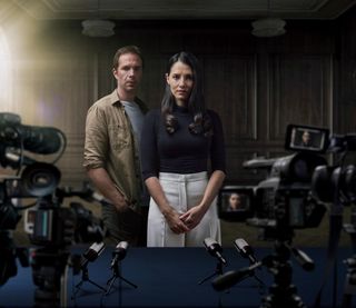Natalie Varga (Charlie Murphy) stands in a wood-panelled room in a black top and white trousers with her hair down. James Alden (James D'Arcy) stands behind her in a khaki shirt, grey t-shirt and jeans. In the foreground we see multiple television cameras, just out of focus, with Natalie's face visible in the viewfinders