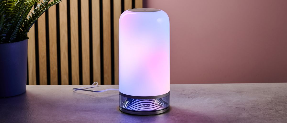 The Govee Table Lamp 2 is lit up with pink and purple colors and sits on a stone surface with wooden slats and a pink wall behind.