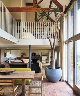 double-height combined living dining space with stone floors and mezzanine with exposed beam and wooden dining table and large indoor plant