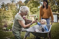 'Sandwich generation' woman pushing a wheelbarrow with a child in a garden next to her older mother