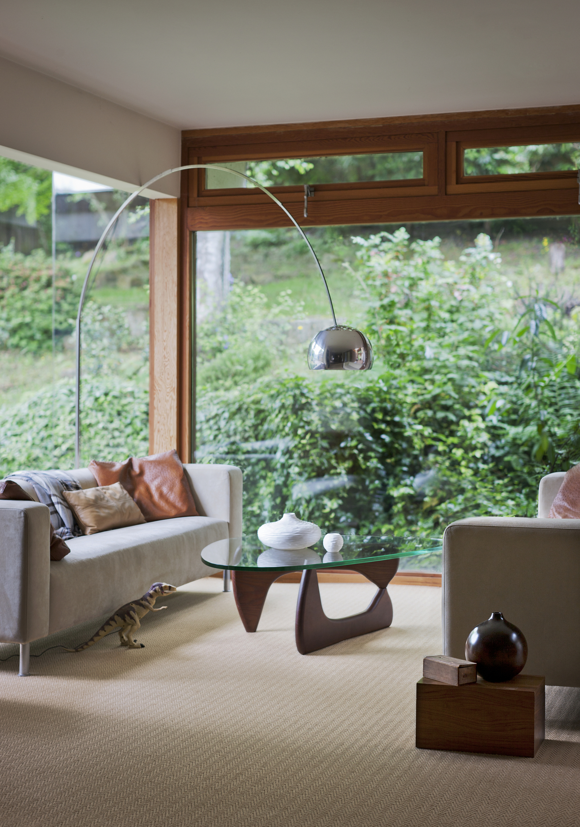 modern living room with beige carpets, gray sofa with cushions, a coffee table, arc lamp leaning over the sofa, and floor to ceiling windows looking to the garden outside