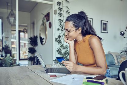 A woman is sitting at a dining table with a laptop, a credit card, and financial statements.