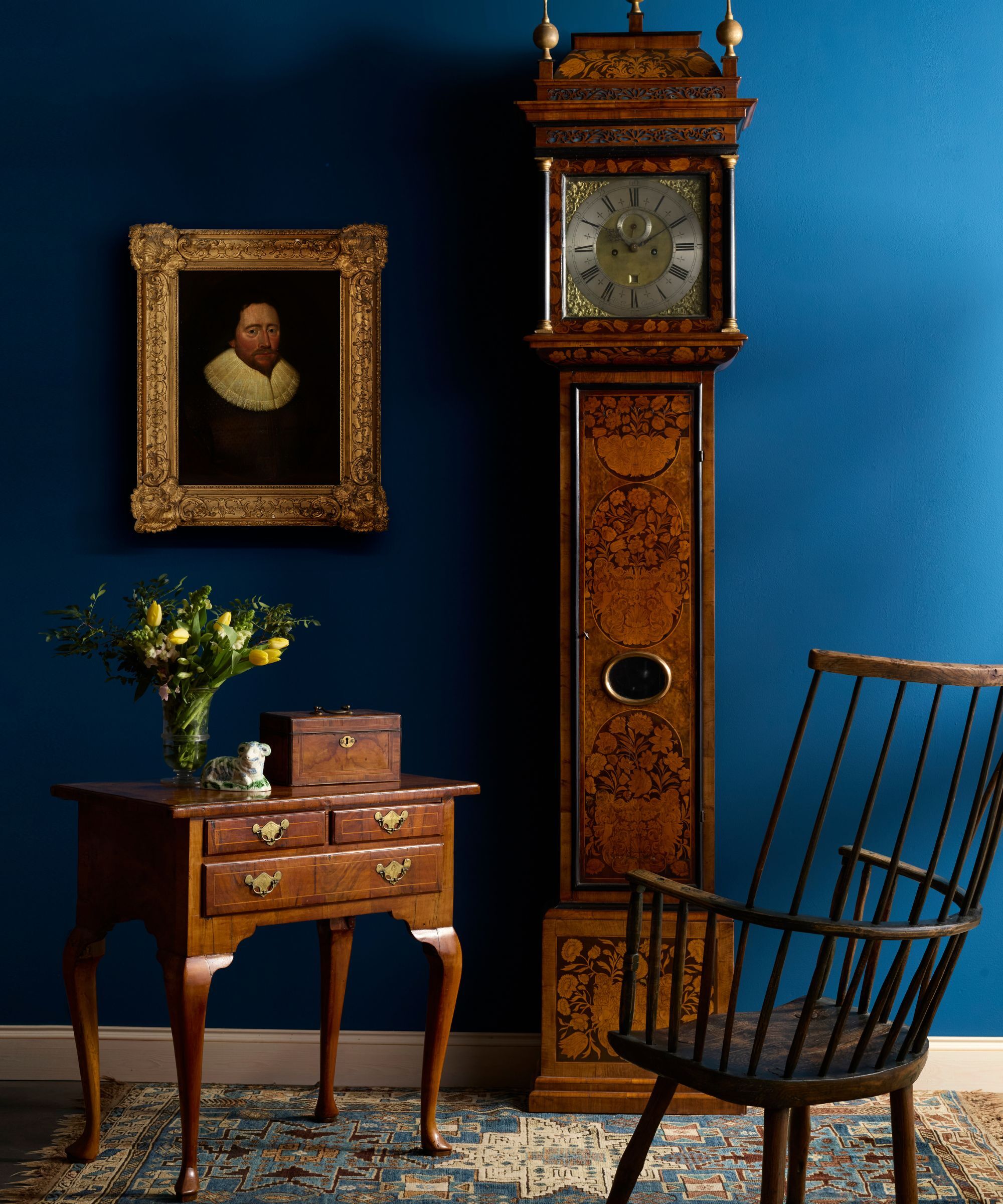A grandfather clock beside an antique chair and desk. On the desk is a glass vase of yellow tulips and foliage. Next to it is a small sculpture and a mahogany box. On the wall behind is a portrait painting in a gold guilt frame. The wall is a bright shade of navy blue.