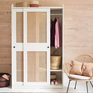 White freestanding wardrobe with rattan inserts, next to a rattan chair