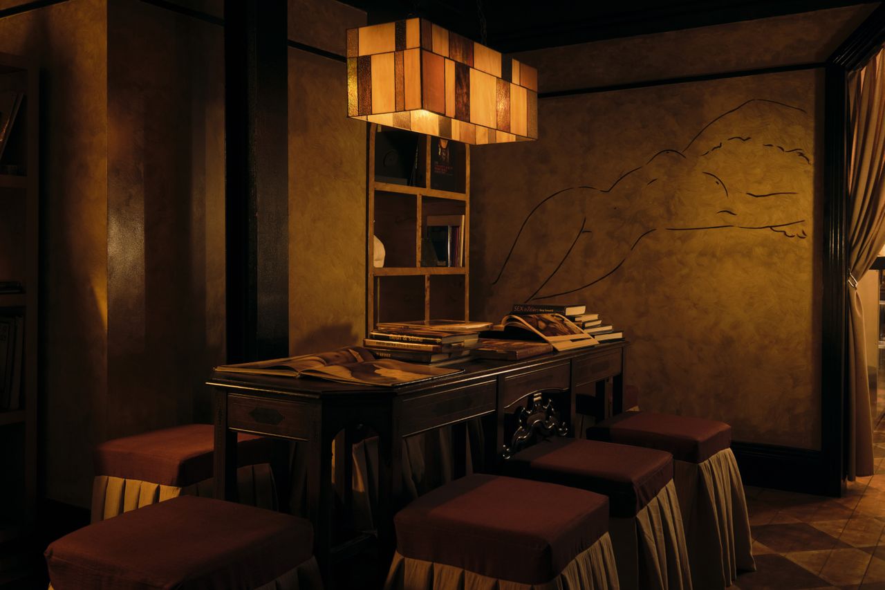 An amber-lit nightclub features retro-inspired furniture echoing the atmosphere of the 20th century, marble busts, plush fabrics, and wall murals.