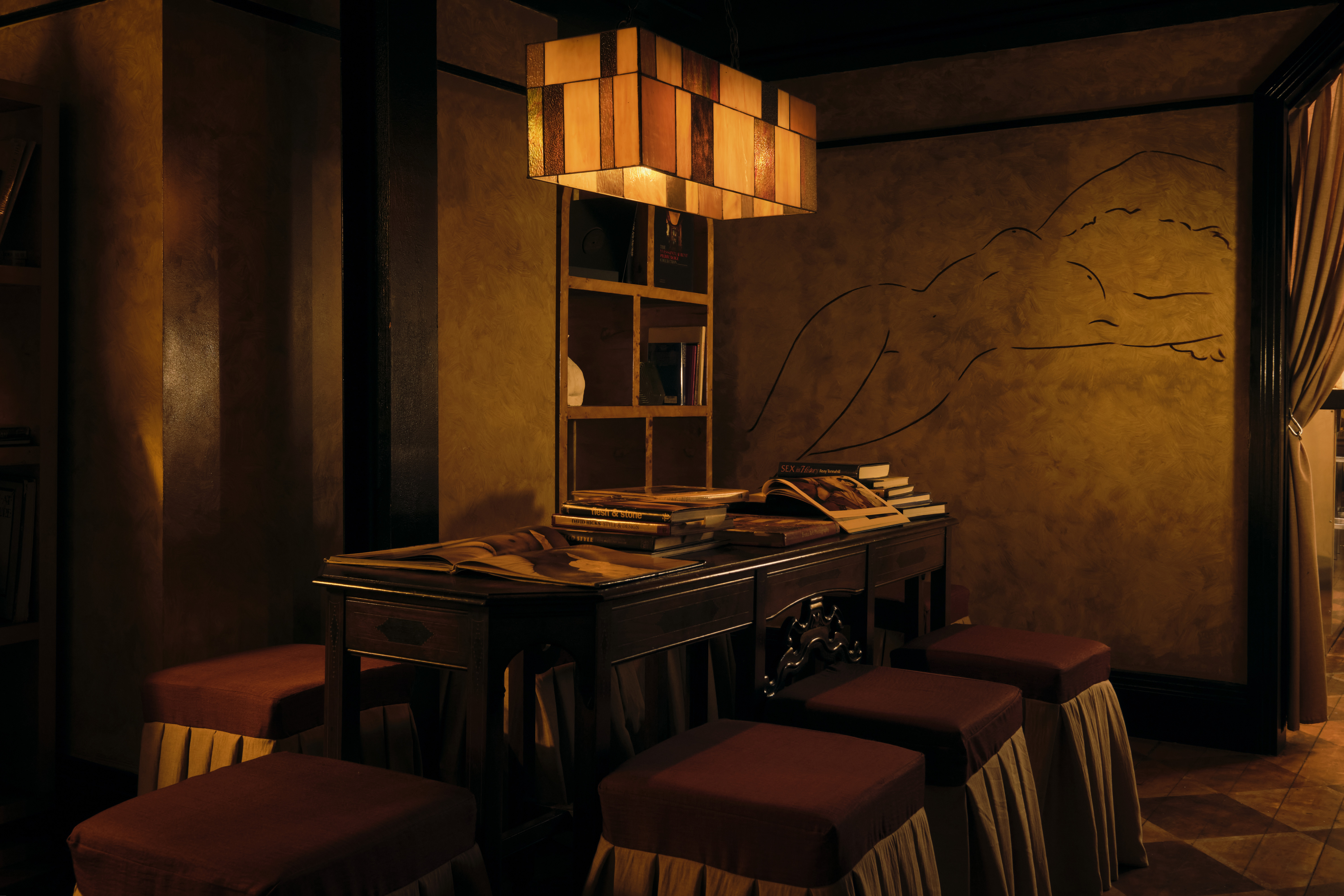 An amber-lit nightclub features retro-inspired furniture echoing the atmosphere of the 20th century, marble busts, plush fabrics, and wall murals.