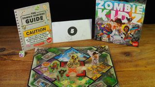 Zombie Kidz Evolution board, box, cards, dice, tokens, and envelopes on a wooden table against a black background