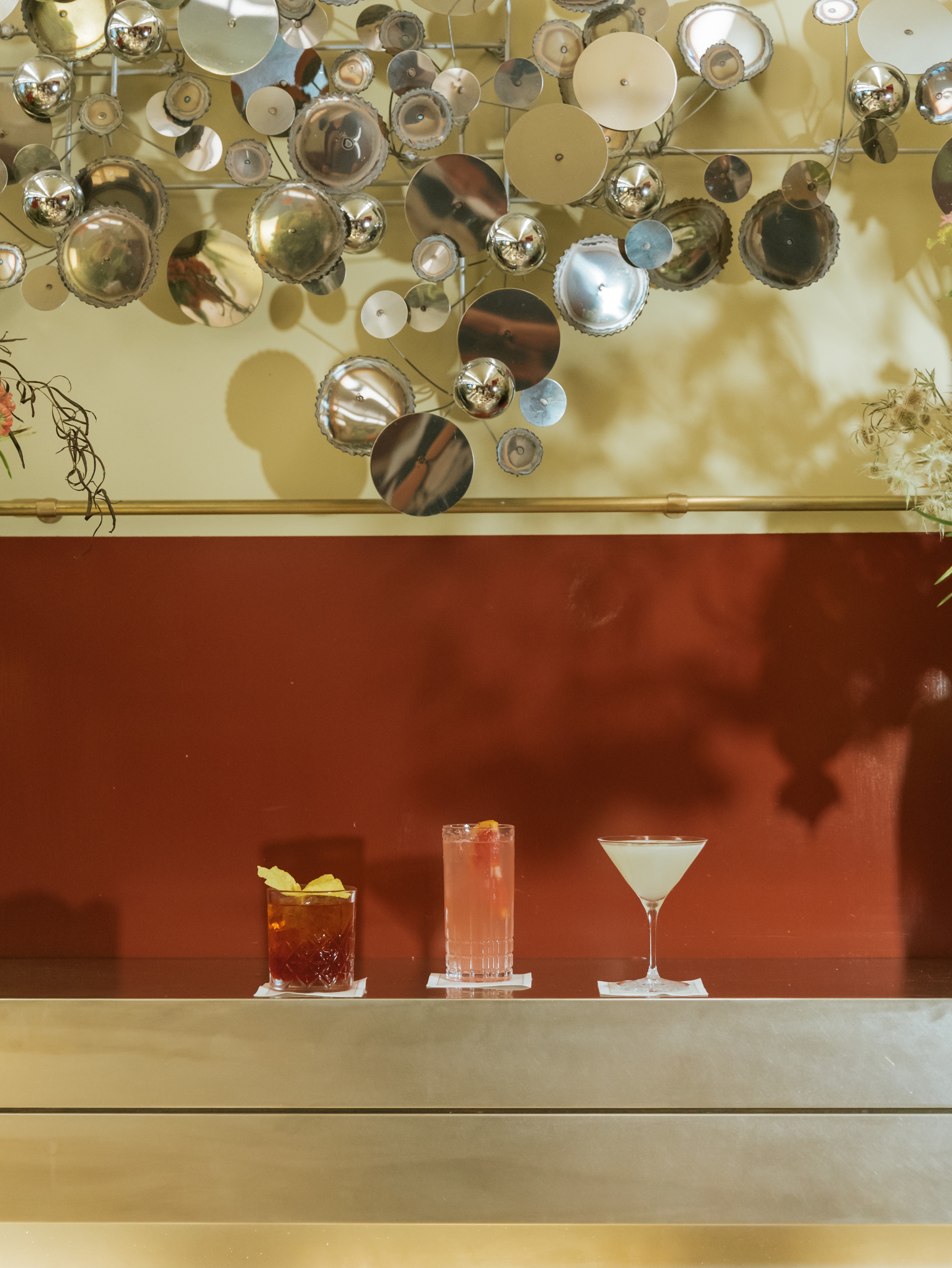 A still life image of a series of high-end cocktails served in thumblers and Martini glasses and sitting atop a chrome surface in a red and pale yellow-tinted restaurant with chrome decorations.