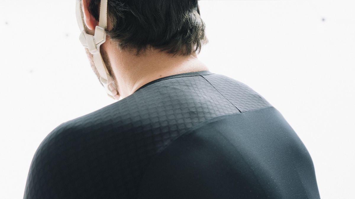 A white man in a tight black cycling jersey stands against a white wall