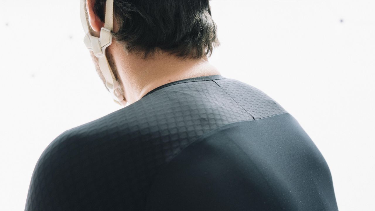 A white man in a tight black cycling jersey stands against a white wall