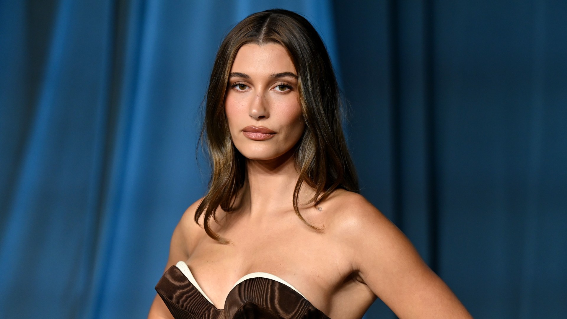 a woman with dark hair posing for paparazzi in a brown strapless gown at a red carpet event