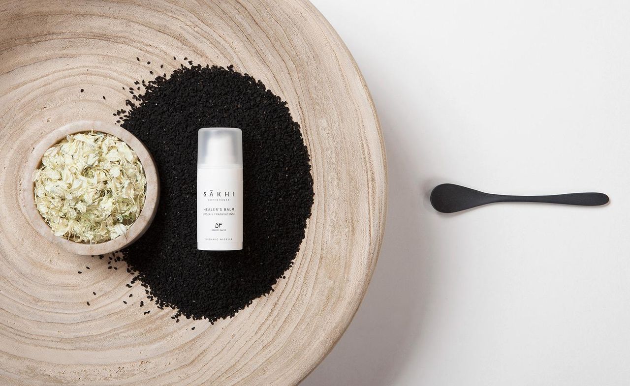 Sakhi healer&#039;s balm bottle in al white, placed on a plate with black seeds and herbs in a bowl, with a black spoon next to it.