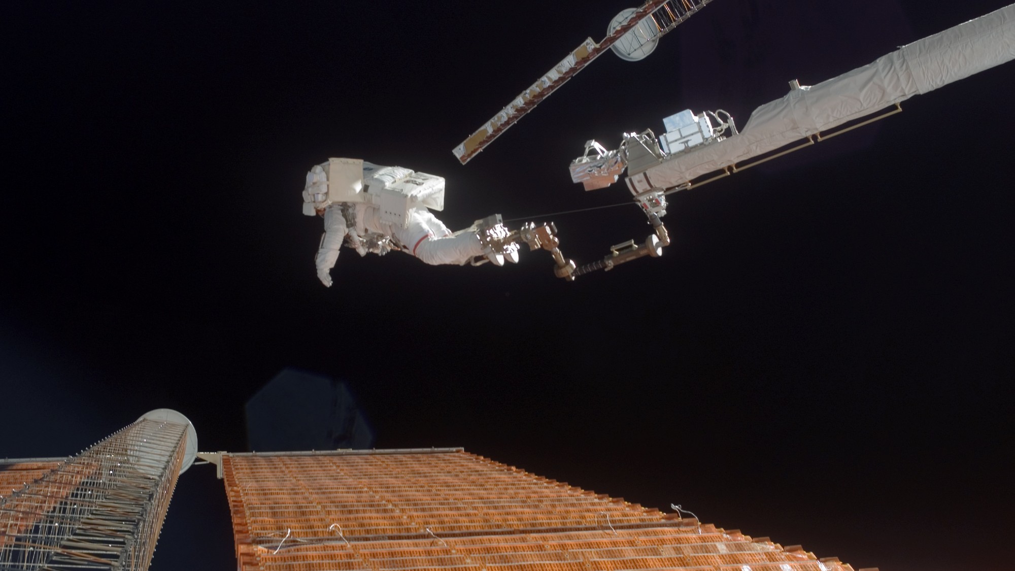 Um astronauta vestindo um volumoso traje espacial branco senta-se na extremidade de um braço robótico acima de um grande painel solar retangular