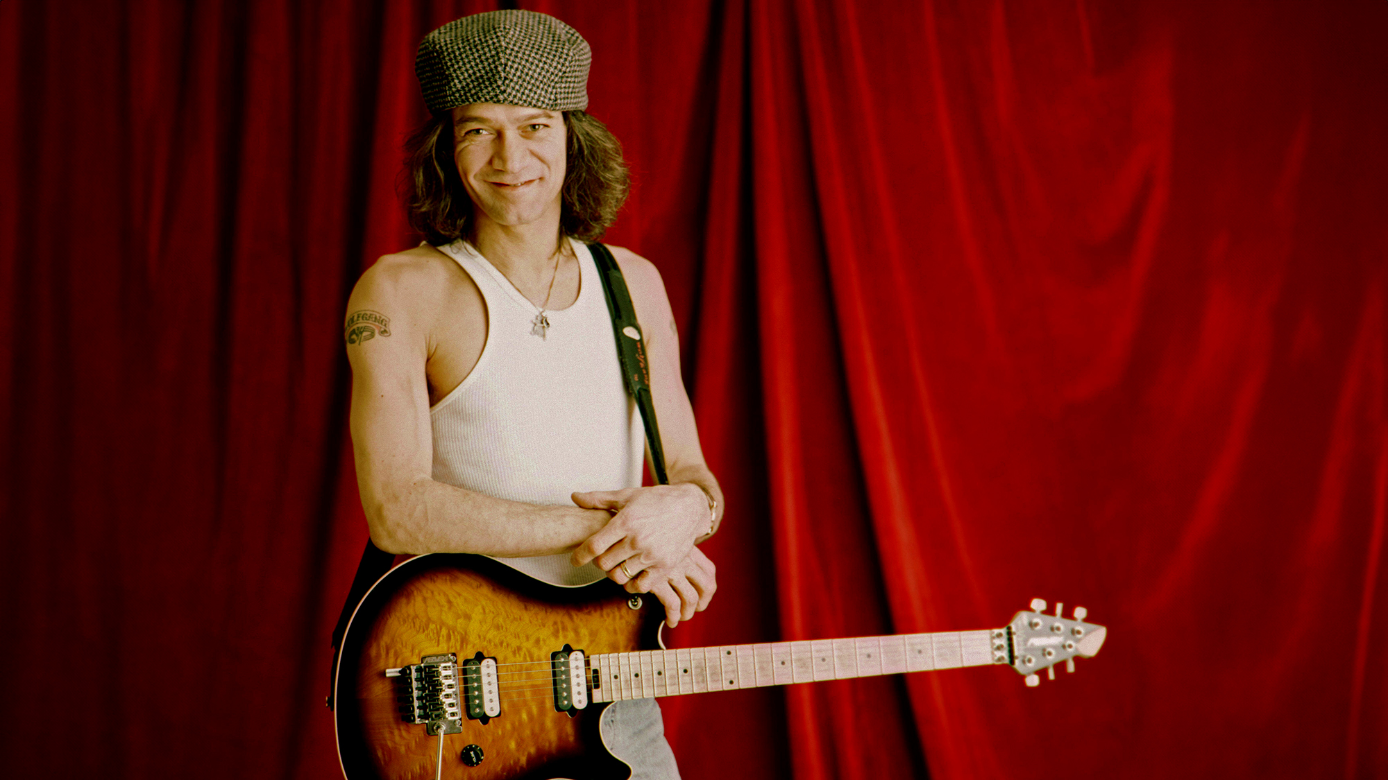 Eddie Van Halen photographed in 1995 posing with an electric guitar in front of a red curtain