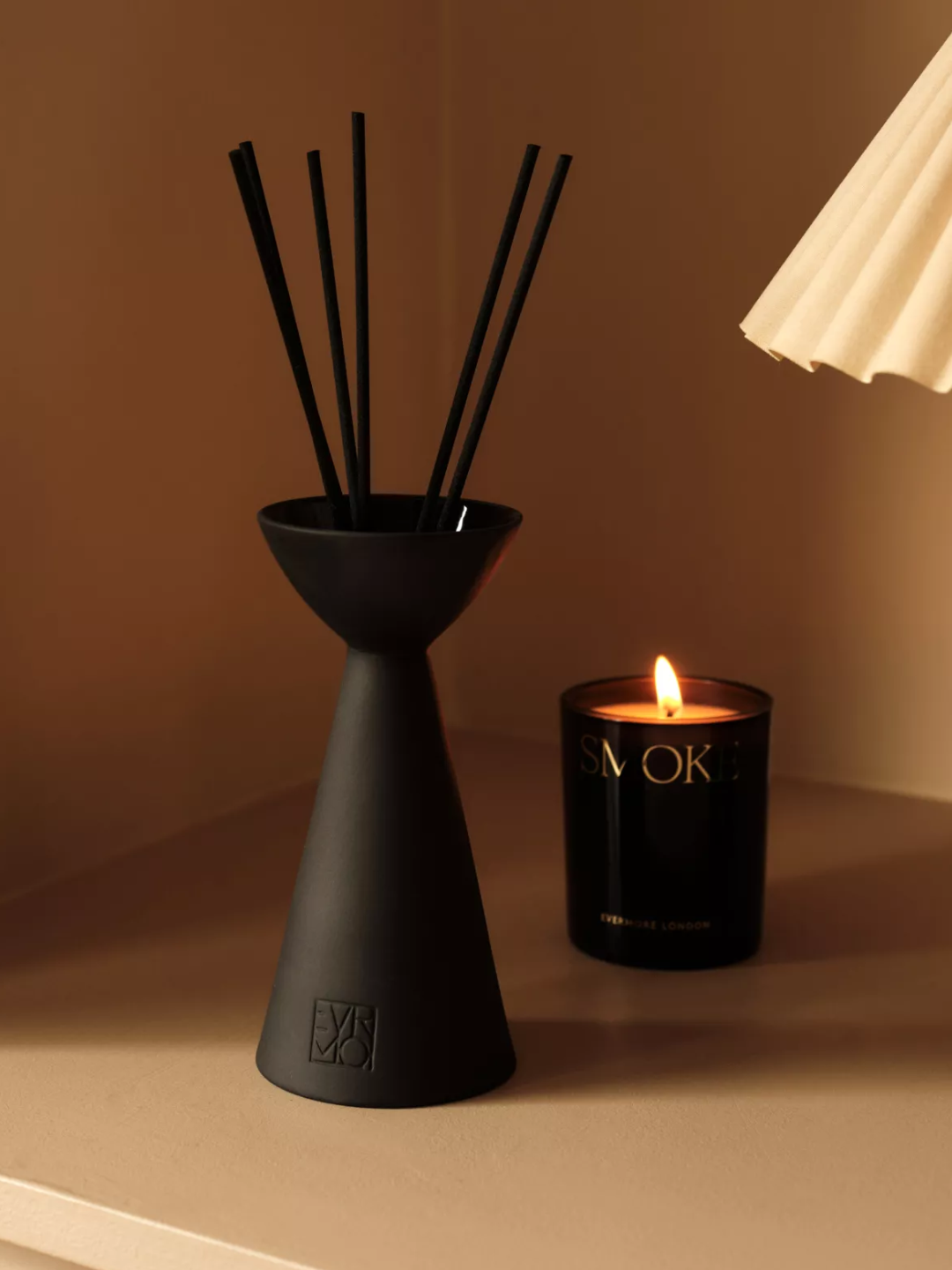 A structural reed diffuser with a handful of black reeds from Evermore beside a lit candle and a quilted lamp shade