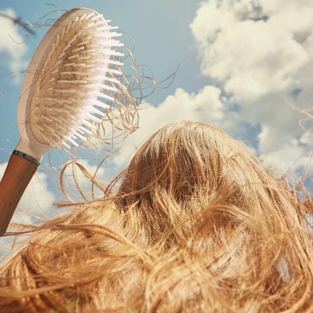 the back of a woman&#039;s head with light brown hair against a blue cloud sky with a hairbrush full of loose hair