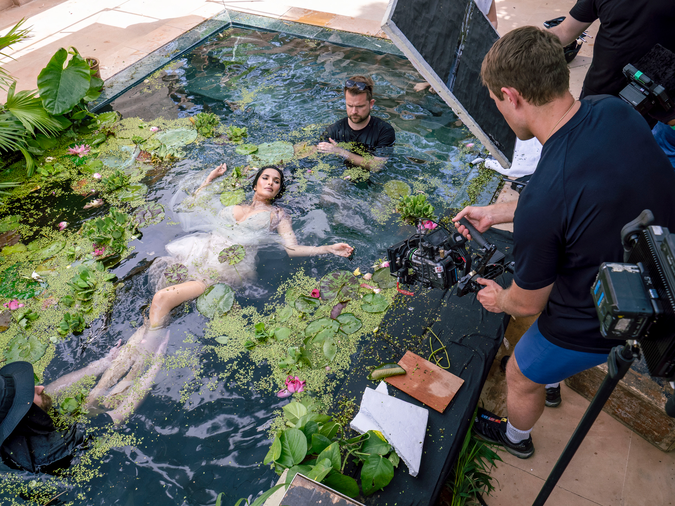 A woman lies in a lake, wearing a silver dress, with a video and photo crew around her, holding cameras and diffusers
