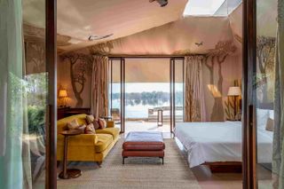 RAAS Chhatrasagar hotel room, yellow settee and side table, white dressed bed to the right, brown ottoman table in the centre, tree illustration on the neutral walls and ceiling, skylight, lamps on side tables, view of the surrounding landscape through the open balcony doors