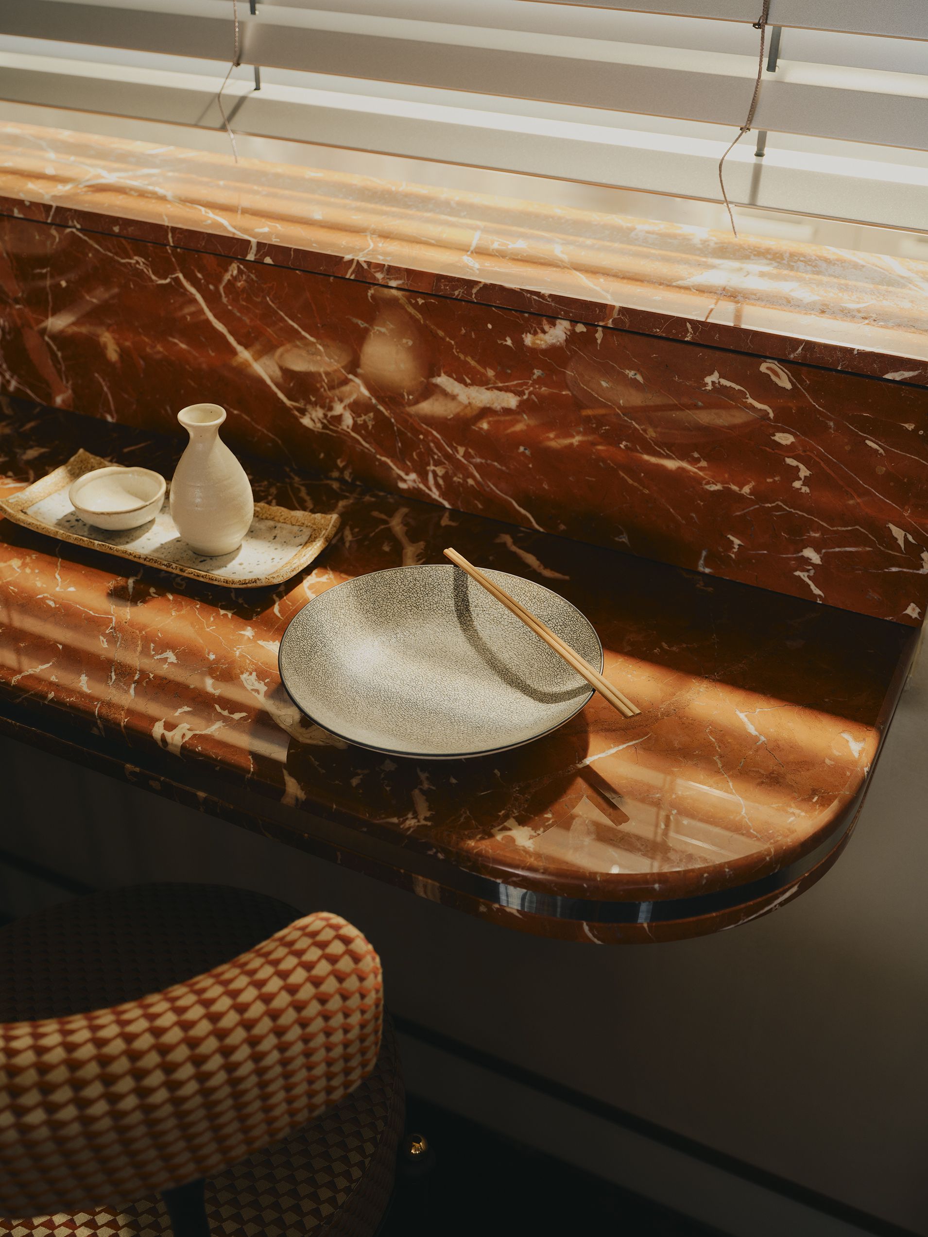 A slim wall-hung marble restaurant table with curved patterned seating with an empty gray plate with a set of chopsticks on top and a tray with a vase and bowl in it for condiments