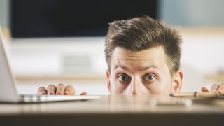 Surprised businessman hiding behind blurry wooden desktop with laptop and other items