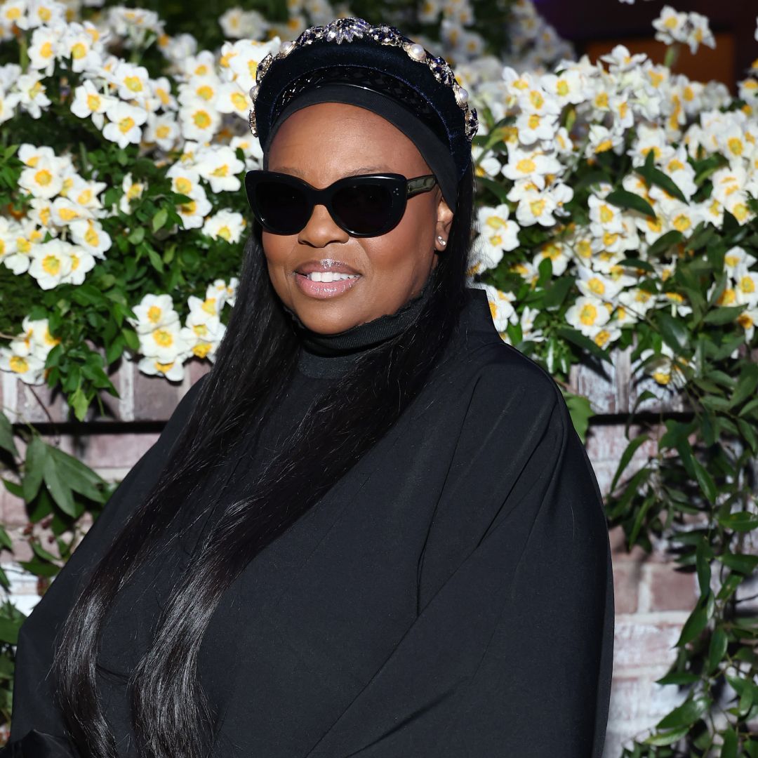 Dame Pat McGrath wearing sunglasses and a black turtleneck sweater. She is standing in front of a bouquet of flowers.