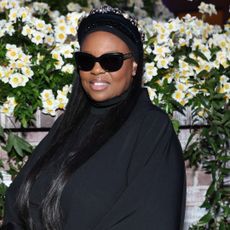 Dame Pat McGrath wearing sunglasses and a black turtleneck sweater. She is standing in front of a bouquet of flowers.